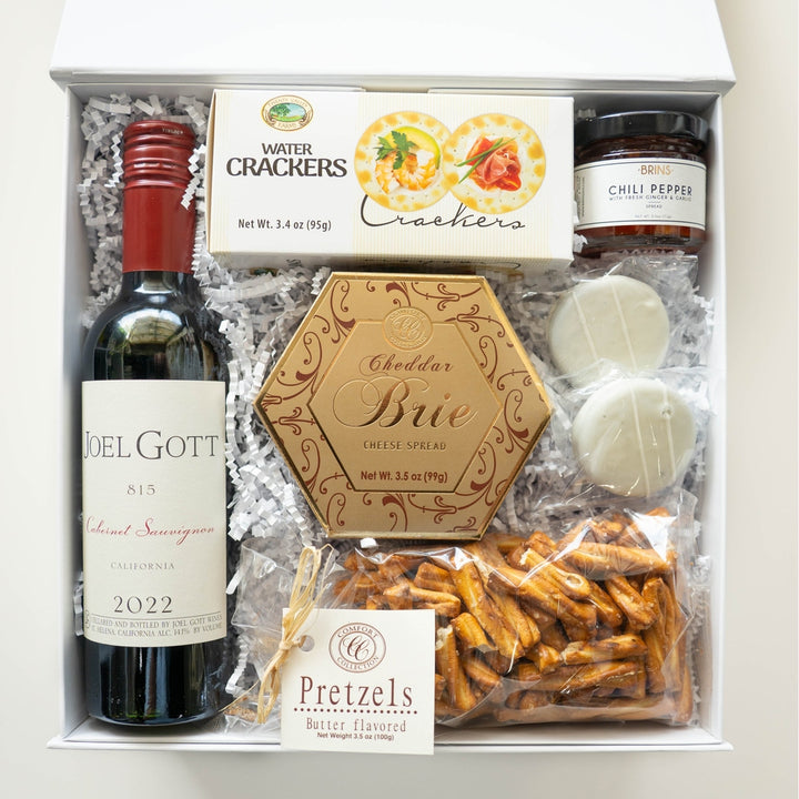 Studio photograph of a Joel Gott wine and cheese gift box featuring a red-capped bottle of Cabernet Sauvignon, a gold hexagonal cheddar brie container, and Valley Farms water crackers nestled in white crinkle-cut paper