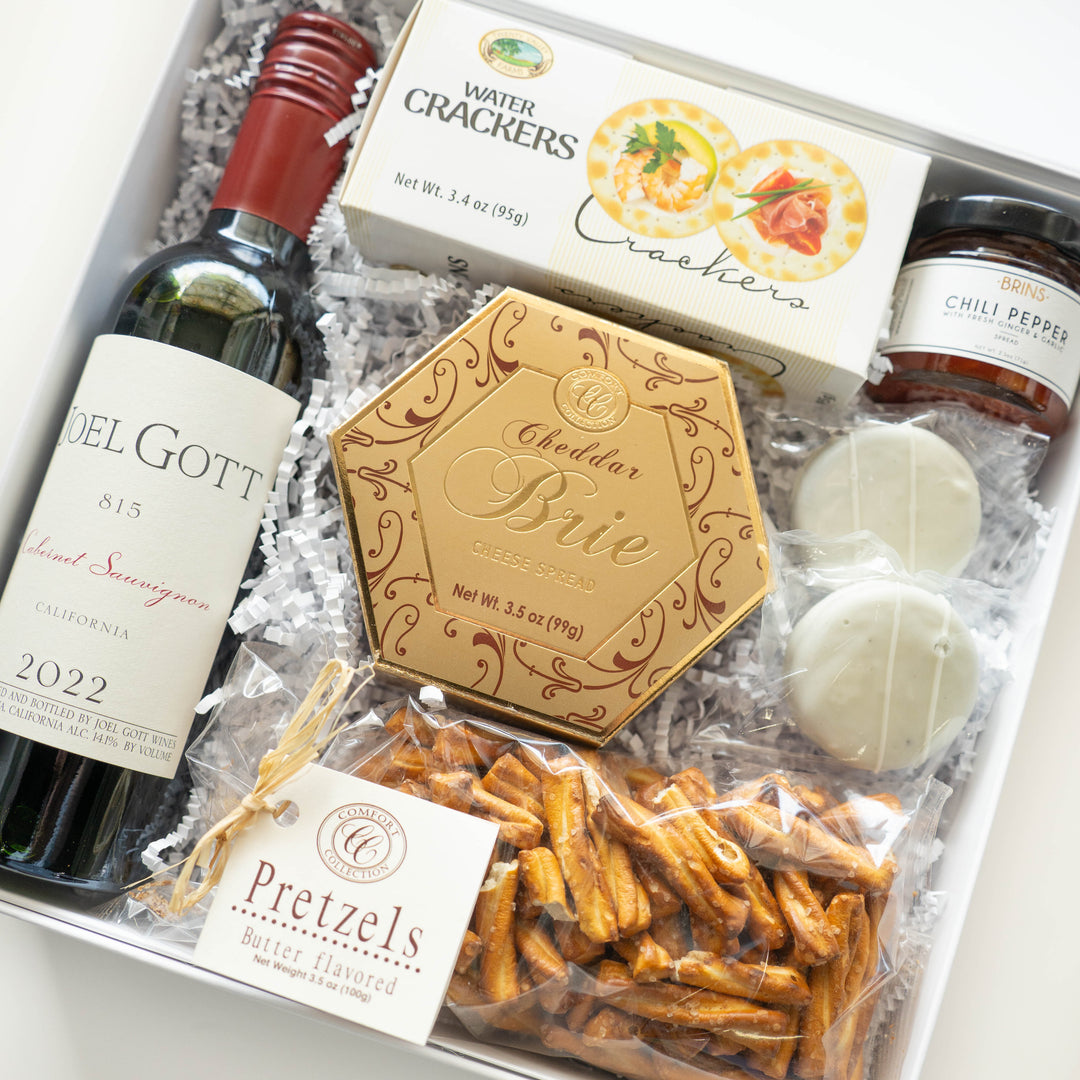 Commercial photograph of a gourmet wine gift box inside a white container featuring a bottle of Joel Gott 2022 Cabernet Sauvignon with a red cap, a gold hexagonal box of Cheddar Brie spread, and a bag of salty pretzels on white shredded paper