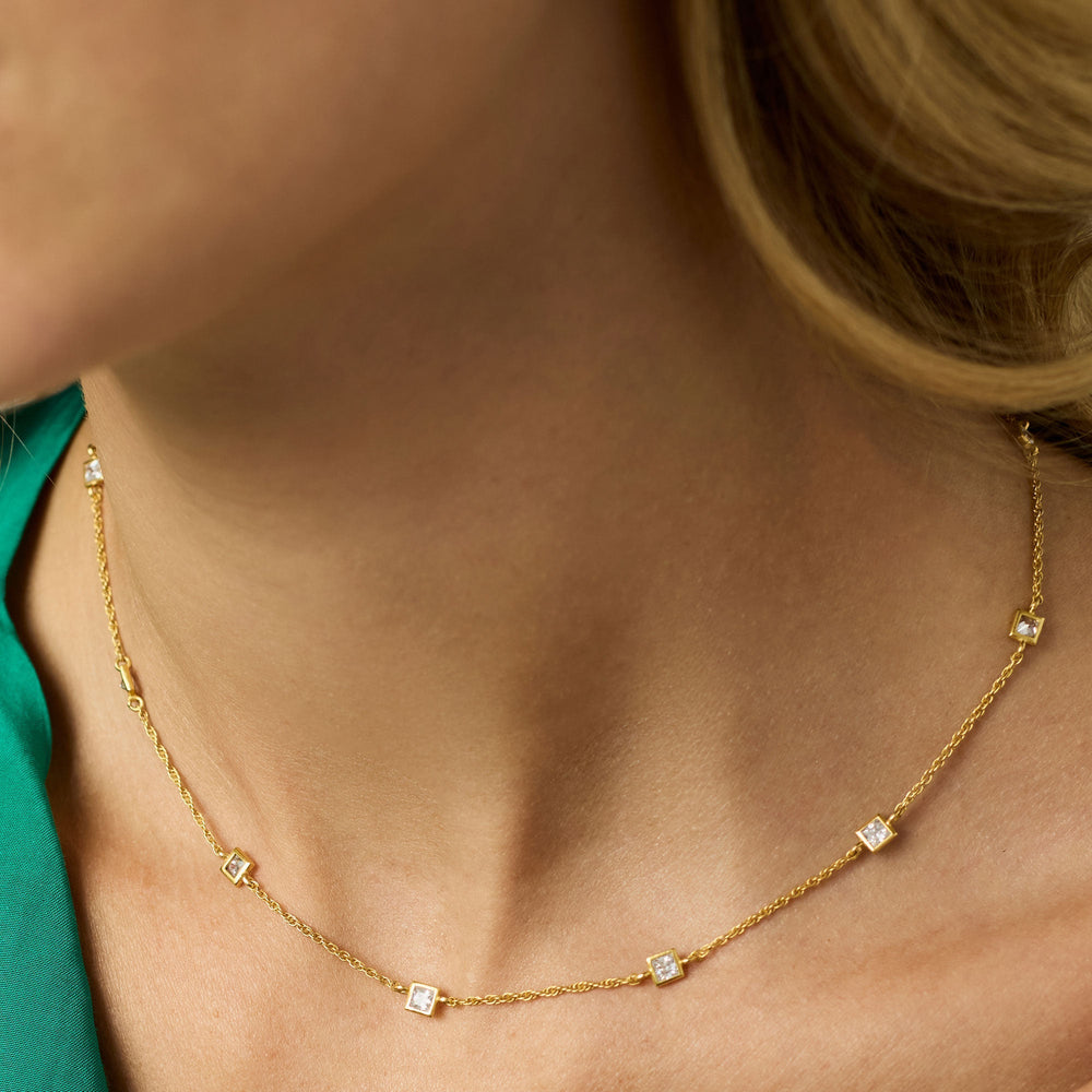 Close-up photograph of a dainty gold station necklace with five square-cut clear stones draped over a woman's collarbone featuring a twisted rope chain and a vibrant green silk blouse