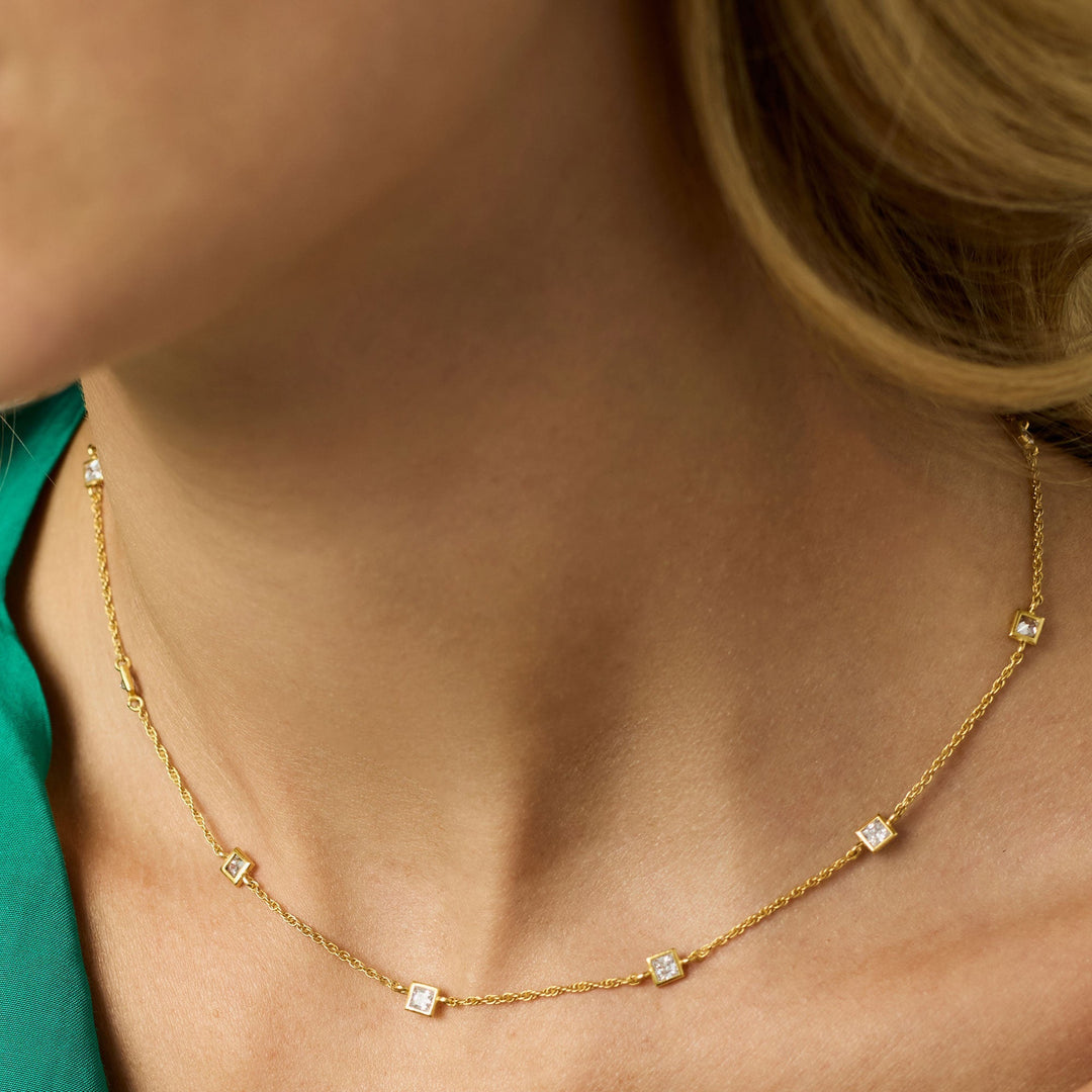 Close-up photograph of a dainty gold station necklace with five square-cut clear stones draped over a woman&