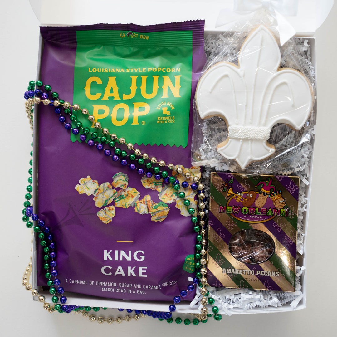 Photograph of a New Orleans Mardi Gras gift box showcasing a purple bag of Cajun Pop popcorn, an iconic white-iced fleur-de-lis sugar cookie, and metallic purple, green, and gold beads