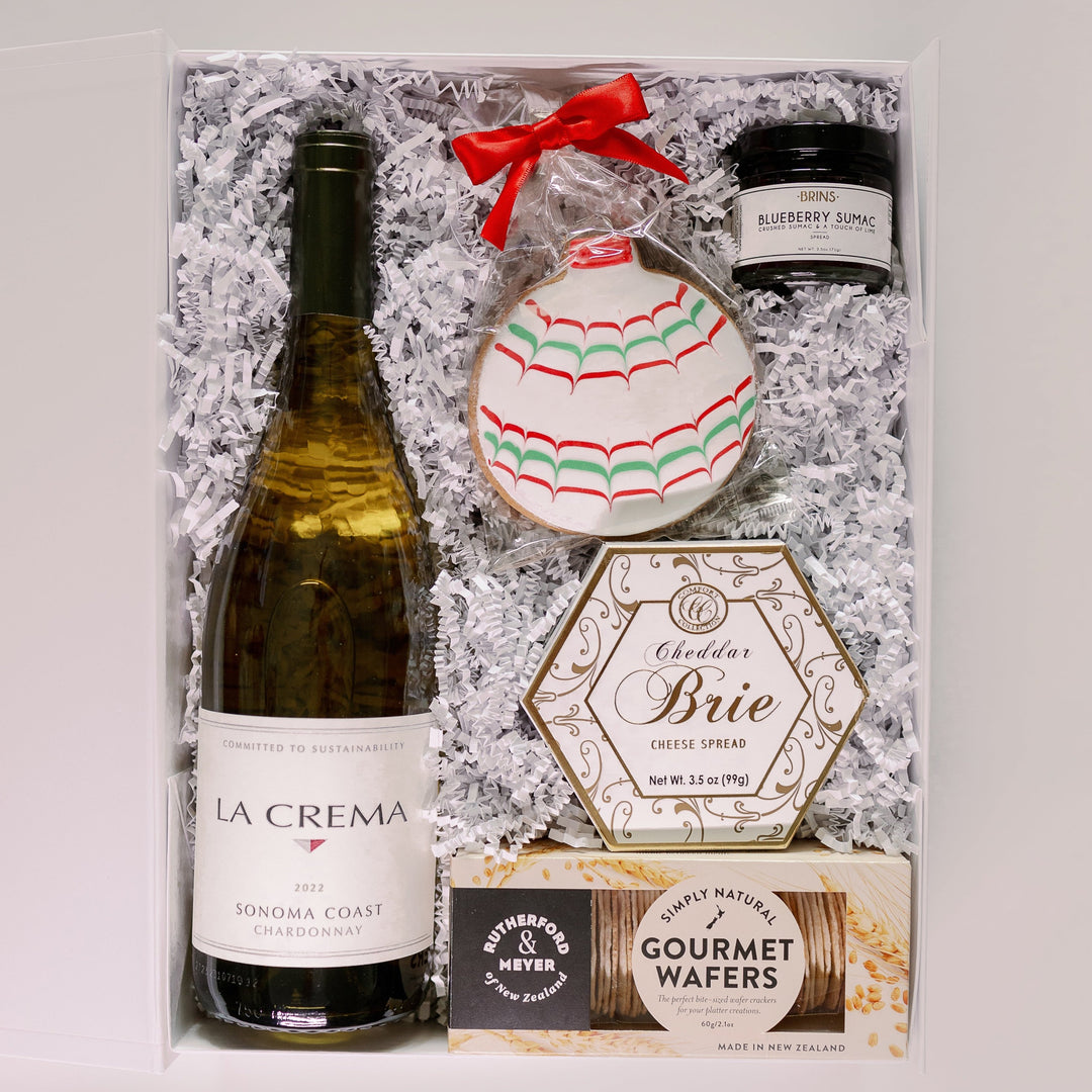 Photograph of a curated holiday gift box featuring a bottle of La Crema Sonoma Coast Chardonnay, a festive hand-iced ornament cookie with a bright red bow, and a hexagonal box of cheddar brie spread nestled in white shredded paper