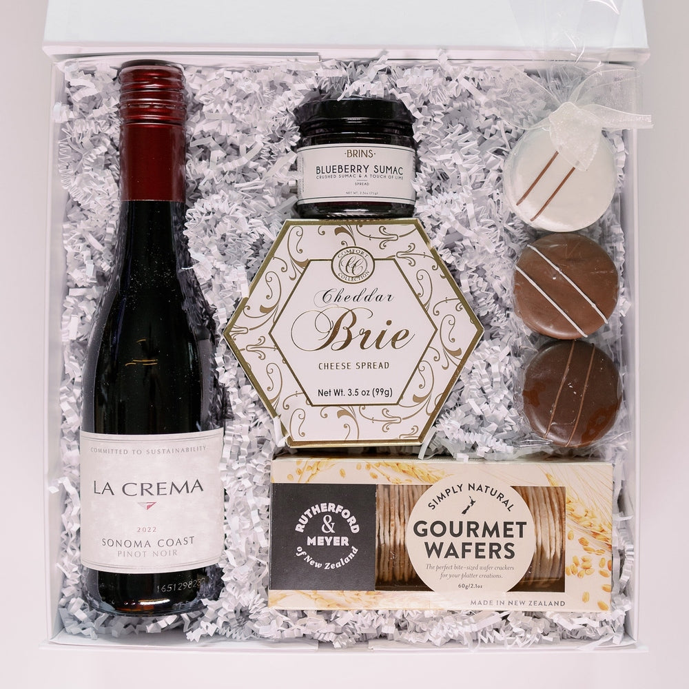 Photograph of a luxury gourmet gift box featuring a bottle of La Crema Sonoma Coast Pinot Noir with a deep red capsule, a gold-patterned hexagonal cheese spread, and three striped chocolate cookies nestled in white shredded paper
