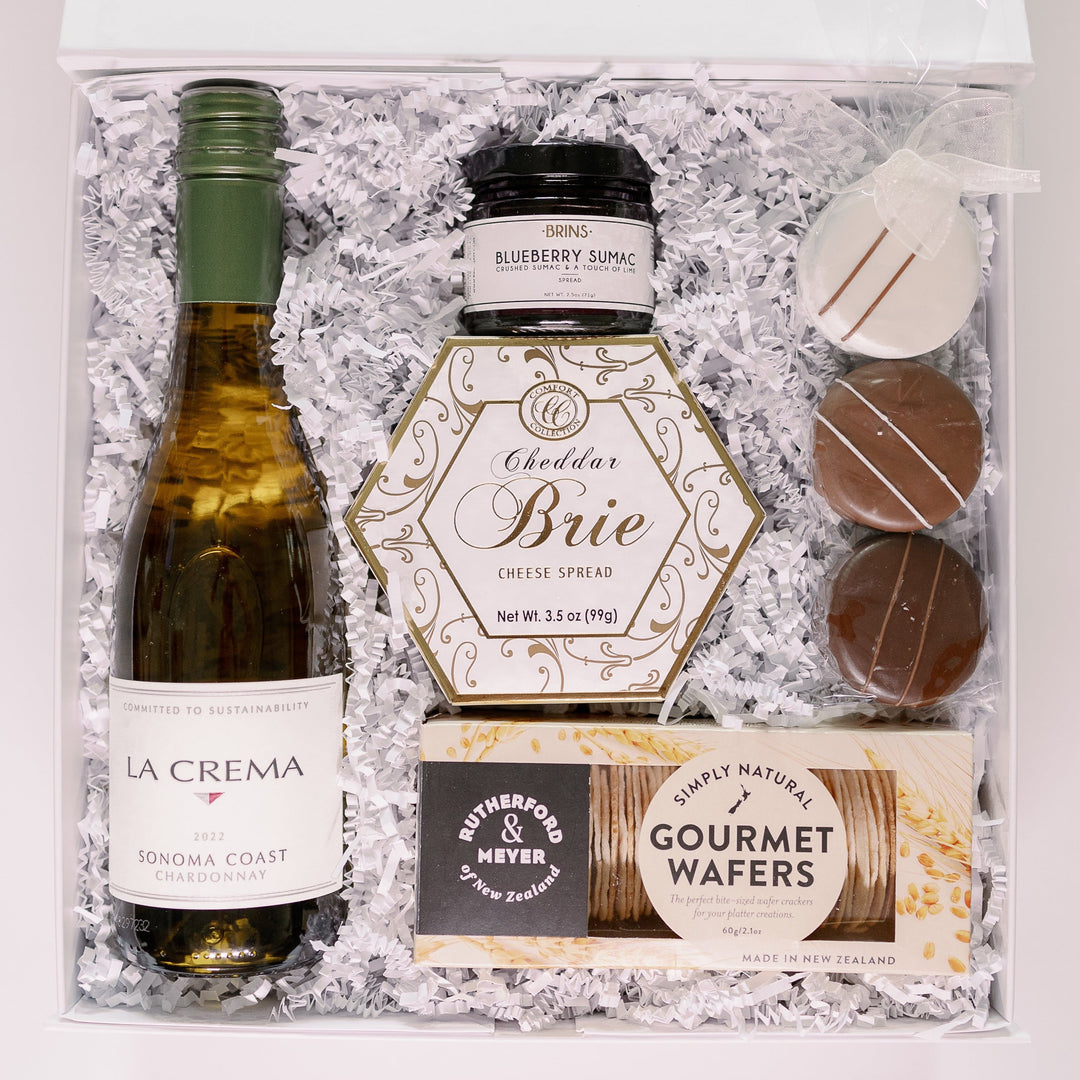 Digital photograph of a white gourmet gift box featuring a bottle of La Crema Sonoma Coast Chardonnay and a gold-trimmed hexagonal box of Cheddar Brie spread nestled in white shredded paper with three chocolate-covered cookies and a box of gourmet wafers