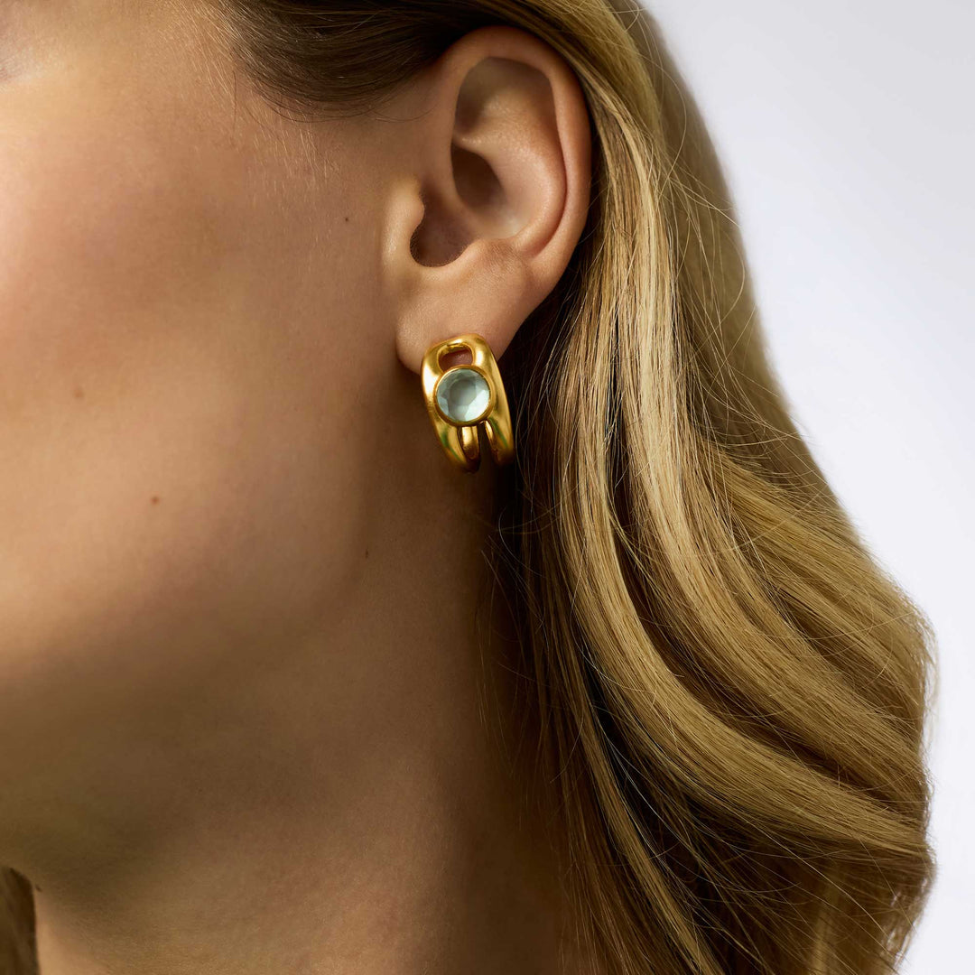 Close-up photograph of a jewelry piece featuring a chunky matte gold hoop earring with a round translucent seafoam green gemstone worn on a model&