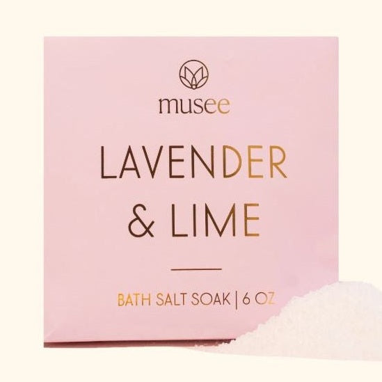 Product photograph by Musee featuring a pink square box of Lavender & Lime bath salt soak with metallic gold lettering and a pile of white crystalline salts on a cream background