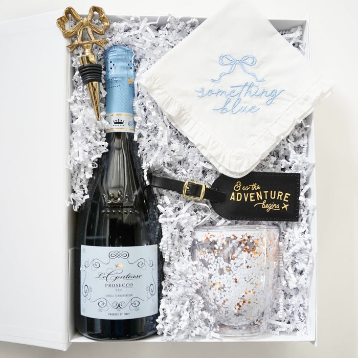 A curated gift box set containing a bottle of Lil' Bordeaux Prosecco, a white embroidered napkin reading 'something blue', a black and gold 'Be the Adventure Queen' tag, and a glittery tumbler, all nestled in white crinkle paper inside an open white box.