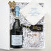 A curated gift box set containing a bottle of Lil' Bordeaux Prosecco, a white embroidered napkin reading 'something blue', a black and gold 'Be the Adventure Queen' tag, and a glittery tumbler, all nestled in white crinkle paper inside an open white box.