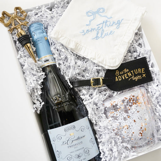A curated gift box containing a bottle of La Cuvée Brut sparkling wine, a white embroidered bandana, a black luggage tag, and a clear glass with confetti, arranged in white crinkle paper.