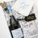 A curated gift box containing a bottle of La Cuvée Brut sparkling wine, a white embroidered bandana, a black luggage tag, and a clear glass with confetti, arranged in white crinkle paper.