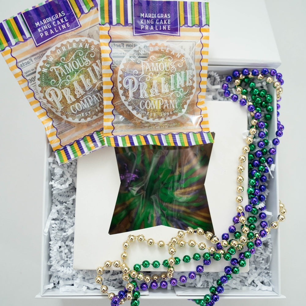 Photograph by New Orleans Famous Praline Company of a Mardi Gras gift box in New Orleans featuring two king cake pralines, a star-shaped window cutout, and vibrant purple, green, and gold beads over white crinkle paper