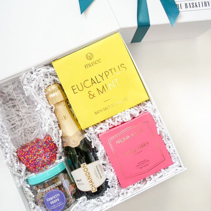 Gift set with a bottle of wine, bath bombs, and a book inside a decorative box with a blue ribbon.