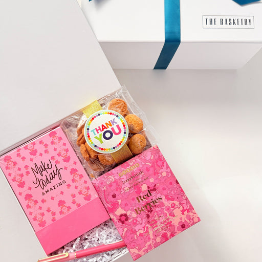 Gift set with pink notebook, cupcake, and cookies in a white box with blue ribbon on a white background.