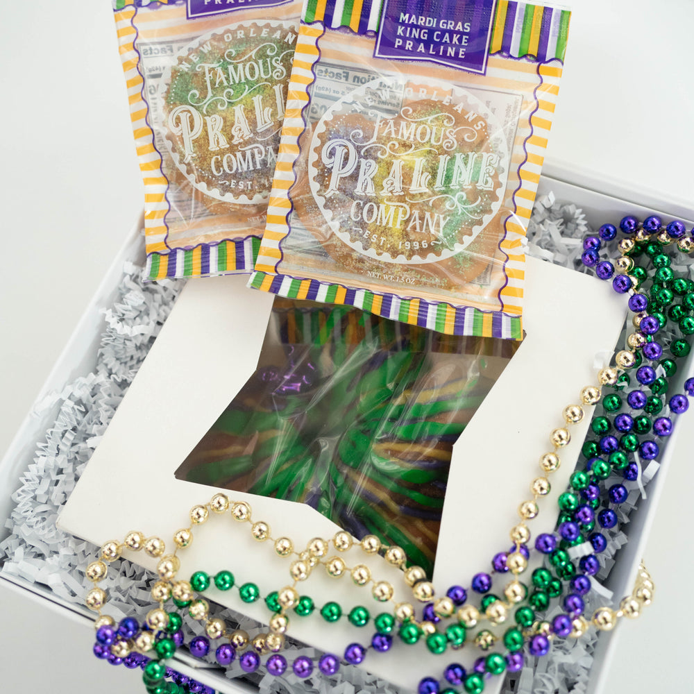 Photograph by New Orleans Famous Praline Company of a Mardi Gras gift box in New Orleans featuring individually wrapped King Cake Pralines and a windowed cake box decorated with vibrant purple, green, and gold beads on white crinkle paper