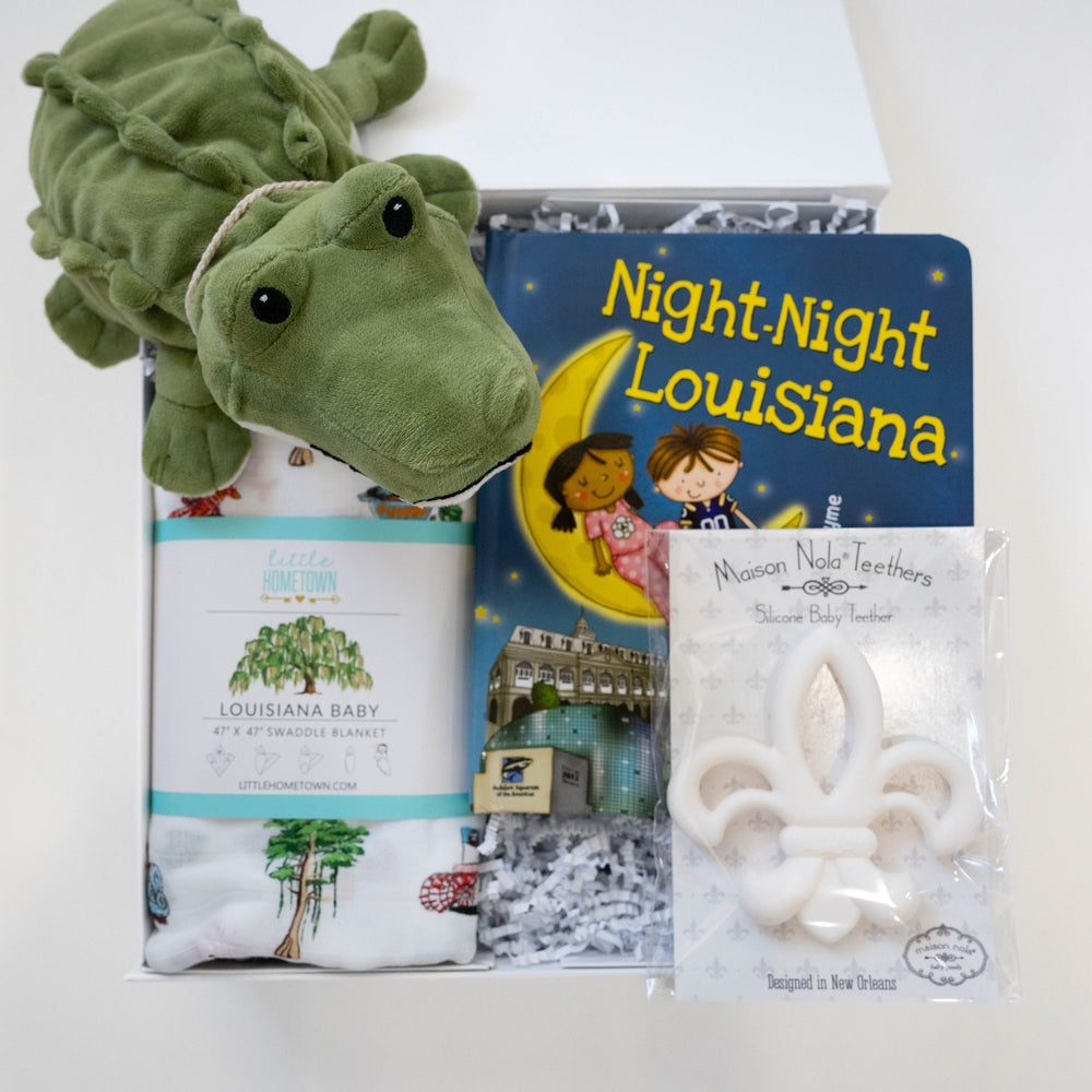 Photograph of a Louisiana-themed baby gift set by Little Hometown and Maison Nola featuring a plush green alligator, a white fleur-de-lis silicone teether, and the Night-Night Louisiana book illustrating the St. Louis Cathedral and Caesars Superdome