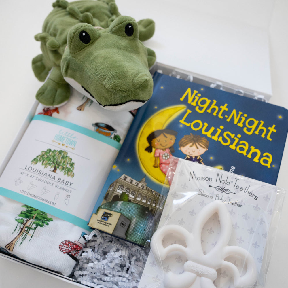 Photograph of a Little Hometown Louisiana baby gift set featuring a plush green alligator, the Night-Night Louisiana book showing the Audubon Aquarium of the Americas, and a white fleur-de-lis silicone teether