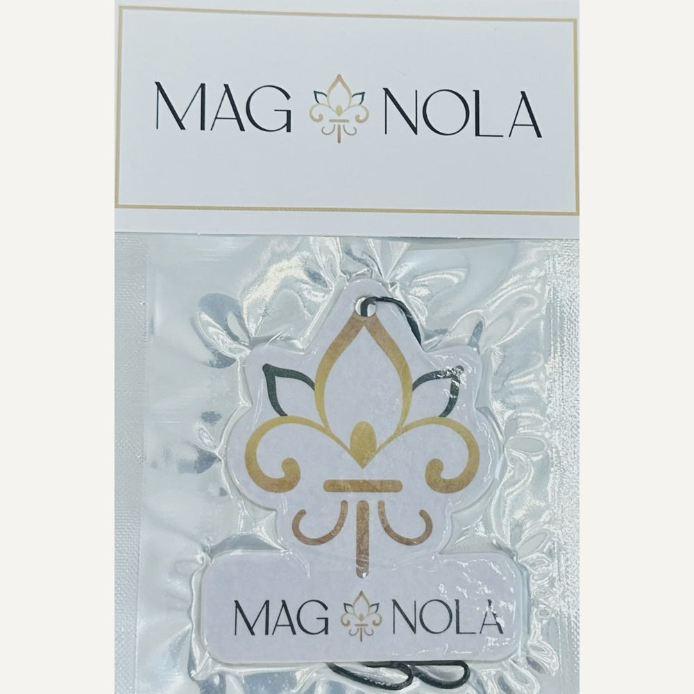 MAG NOLA brand car air freshener featuring a gold and black fleur-de-lis symbol from New Orleans showcased in clear plastic packaging with a white branded header tag
