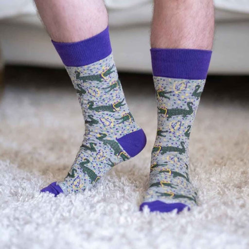 Digital photograph of a person wearing novelty grey crew socks featuring a repeating green alligator and purple bead Mardi Gras pattern with vibrant purple cuffs while standing on a plush white shag rug