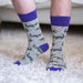 Digital photograph of a person wearing novelty grey crew socks featuring a repeating green alligator and purple bead Mardi Gras pattern with vibrant purple cuffs while standing on a plush white shag rug