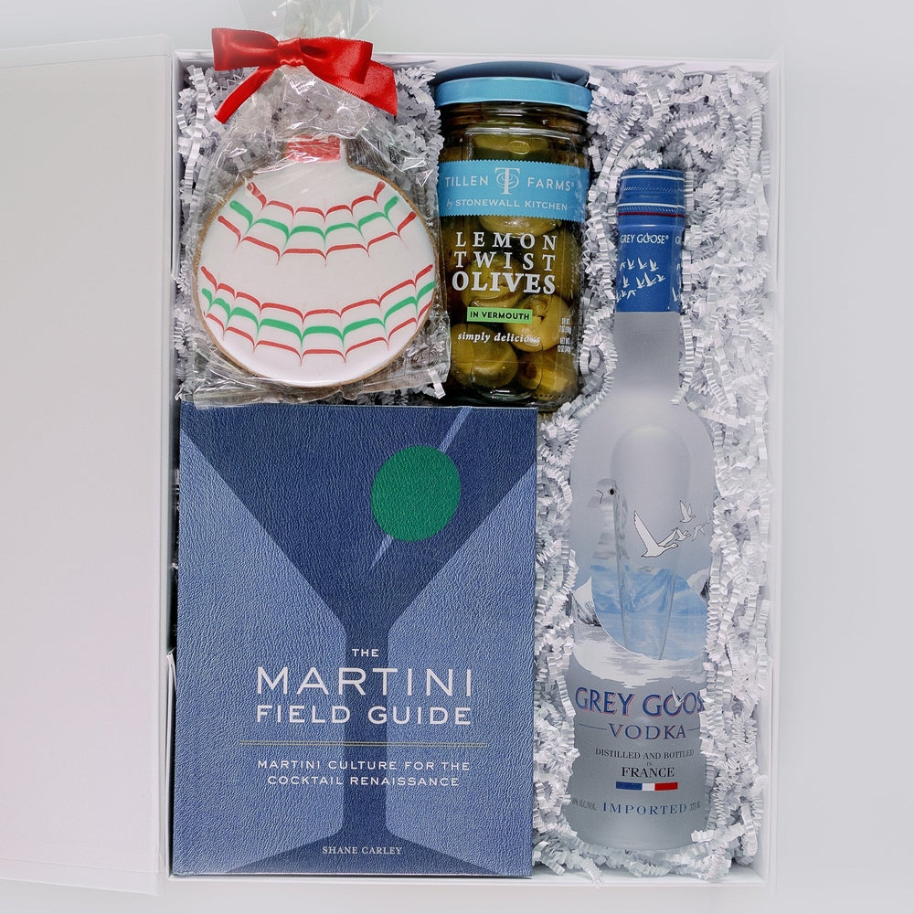 Photograph of a martini-themed gift set in a white box featuring a Grey Goose Vodka bottle, Tillen Farms Lemon Twist Olives, and a blue Martini Field Guide book accompanied by white crinkle paper and a scalloped red and green holiday cookie