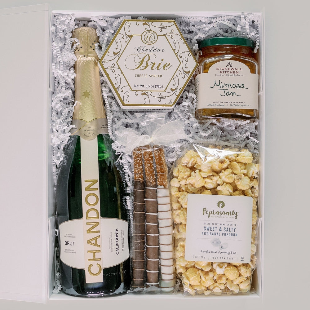 Photograph of a luxury gift box featuring a bottle of Chandon Brut and Stonewall Kitchen Mimosa Jam alongside a hexagonal box of Cheddar Brie cheese, chocolate-covered pretzels with a white bow, and golden artisanal popcorn on white crinkle paper