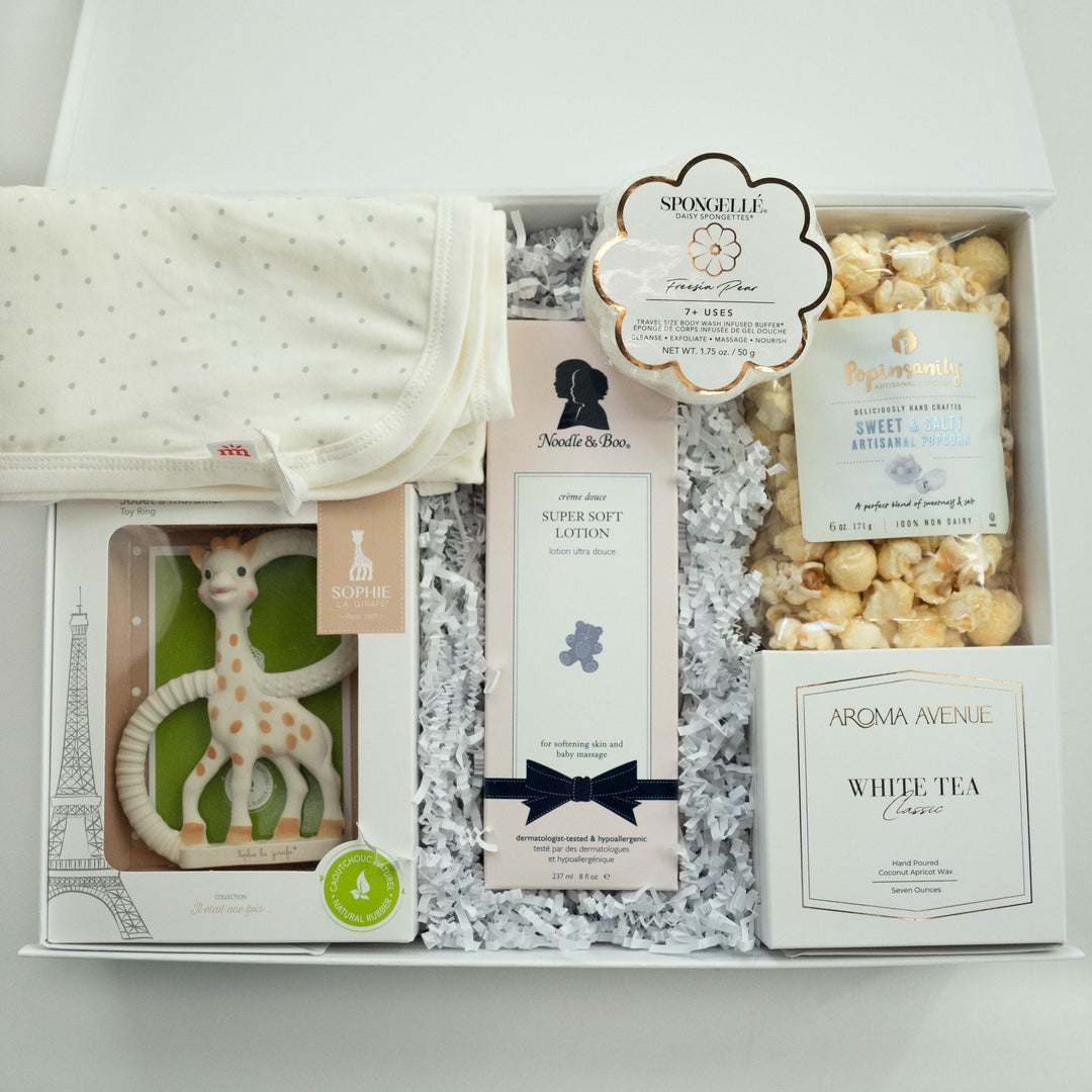 Photograph of a luxury baby gift box containing a Sophie la Girafe teether toy beside an Eiffel Tower illustration, pink Noodle & Boo lotion with a navy bow, and a bag of artisanal popcorn