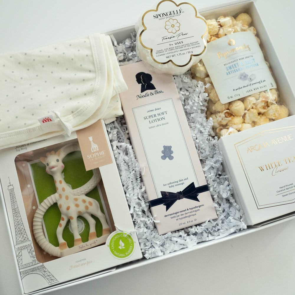 Photograph of a luxury baby gift set containing a Sophie la Girafe teether featuring an Eiffel Tower illustration, a flower-shaped sponge, and a white polka-dot blanket nestled in white crinkle paper