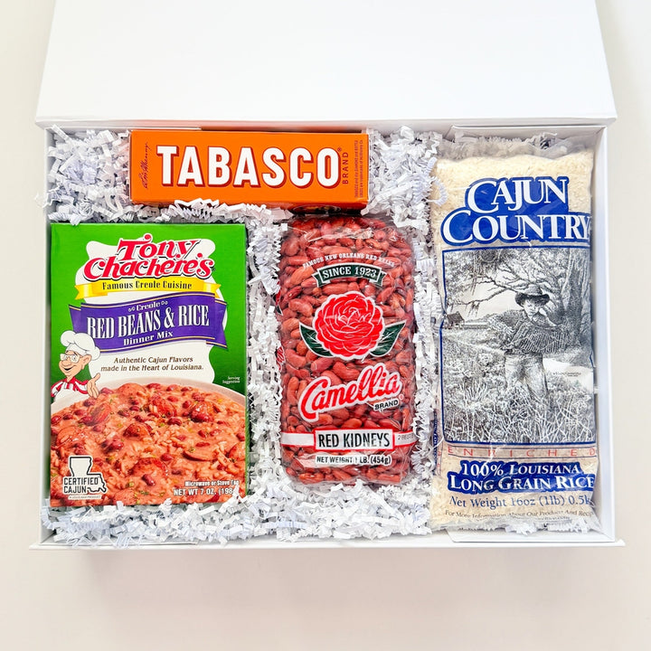 Still life photograph of a Louisiana culinary gift box featuring iconic Tabasco and Tony Chachere’s brands with vibrant orange packaging, red Camellia beans, and blue Cajun Country rice on white crinkle paper