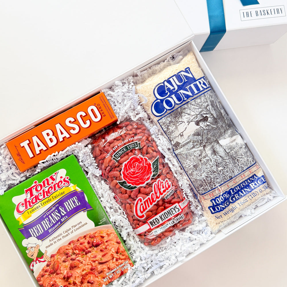 Photograph by The Basketry of a Louisiana culinary gift box featuring iconic brands Tabasco, Tony Chachere’s, and Camellia Red Kidneys with vibrant orange packaging and a blue ribbon on white crinkle paper