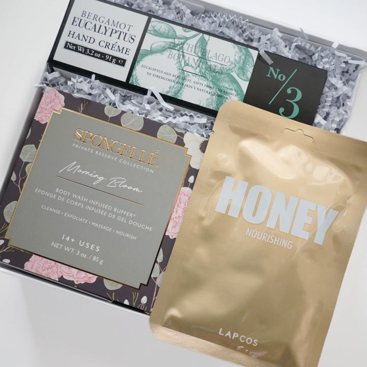 Photograph of a luxury beauty gift box featuring Archipelago Botanicals bergamot eucalyptus hand cream, a floral Spongellé Morning Bloom buffer, and a metallic gold LAPCOS honey mask resting on white crinkle paper
