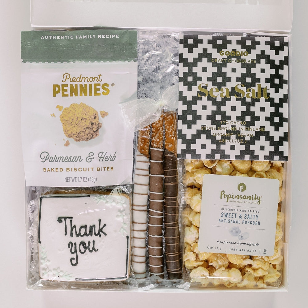 Photograph of a curated gourmet snack gift box featuring a frosted Thank you cookie, chocolate-drizzled pretzel rods tied with a white bow, and a black and white geometric patterned sea salt chocolate bar arranged inside a white container