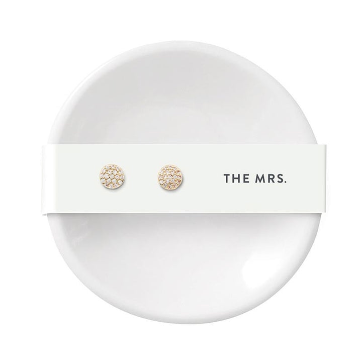 Studio photograph of shimmering gold pave stud earrings mounted on a white display card with black THE MRS text resting atop a minimalist white ceramic ring dish