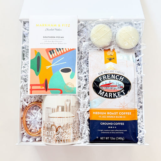 A curated gift box set containing a Mariah & Fitt Southern Pecan box, a bag of French Market ground coffee, two cream macarons, a ceramic mug, and a small gold ring, all arranged in white crinkle paper inside an open white gift box.