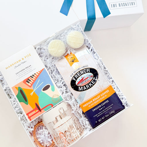 A curated gift box set from The Basketwork, featuring a white gift box with a blue ribbon bow, an open box filled with shreds, a colorful card, a bag of French Market coffee, a glass mug, and two macarons, photographed on a light beige background.