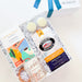 A curated gift box set from The Basketwork, featuring a white gift box with a blue ribbon bow, an open box filled with shreds, a colorful card, a bag of French Market coffee, a glass mug, and two macarons, photographed on a light beige background.