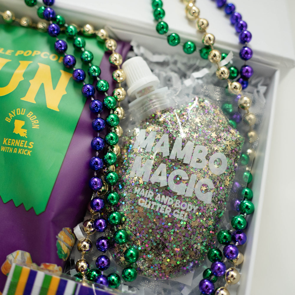 Photograph of a Mardi Gras themed gift box featuring a pouch of Mambo Magic hair and body glitter gel, shimmering purple, green, and gold beads, and a Bayou Born popcorn bag displaying the Louisiana state map icon