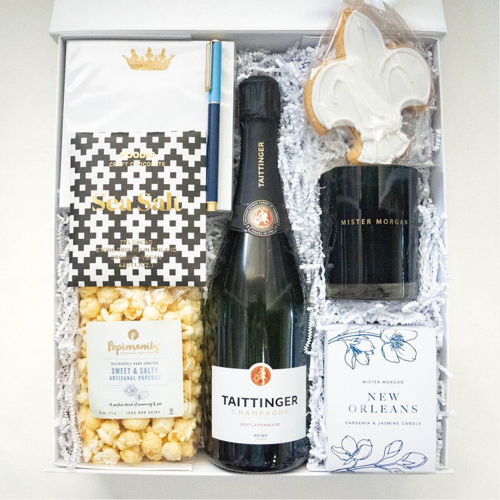 Photograph of a New Orleans themed gift box featuring a bottle of Taittinger Champagne, a white-iced fleur-de-lis cookie, and a gold crown card nestled in white shredded paper