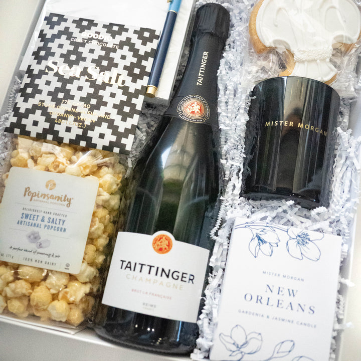 Photograph of a curated luxury New Orleans gift box by Mister Morgan featuring a bottle of Taittinger Champagne, an iconic white-iced fleur-de-lis cookie, and a New Orleans gardenia and jasmine candle alongside black and white geometric sea salt chocolate and artisanal popcorn nestled in white crinkle paper