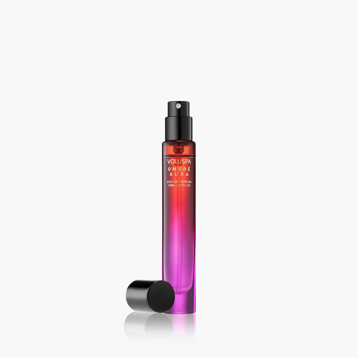 Product photograph by Voluspa of an Ombré Aura Eau de Parfum spray bottle featuring a vibrant red to purple gradient glass vial and a black spray nozzle with its cap resting on a reflective white surface