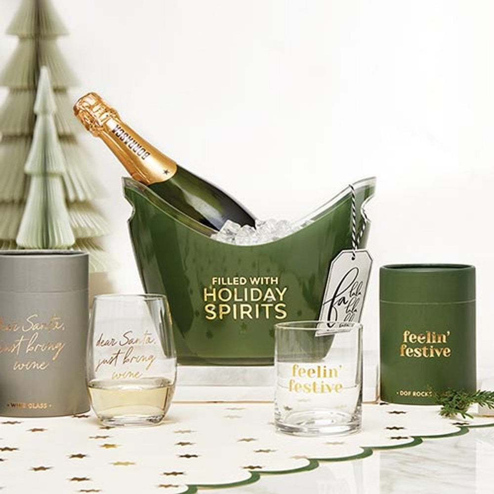 Studio photograph of a festive holiday beverage display on a tabletop featuring a green ice bucket with gold lettering, a Borrasca champagne bottle with gold foil, and a cream-colored paper Christmas tree beside a star-patterned runner