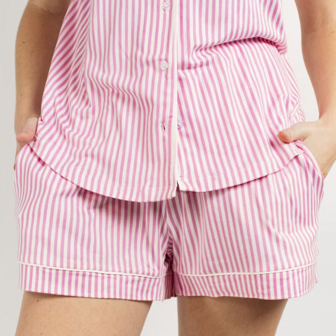 Studio photograph of a person wearing a pink and white vertical striped pajama set featuring a button-down shirt and matching shorts with white piping and side pockets
