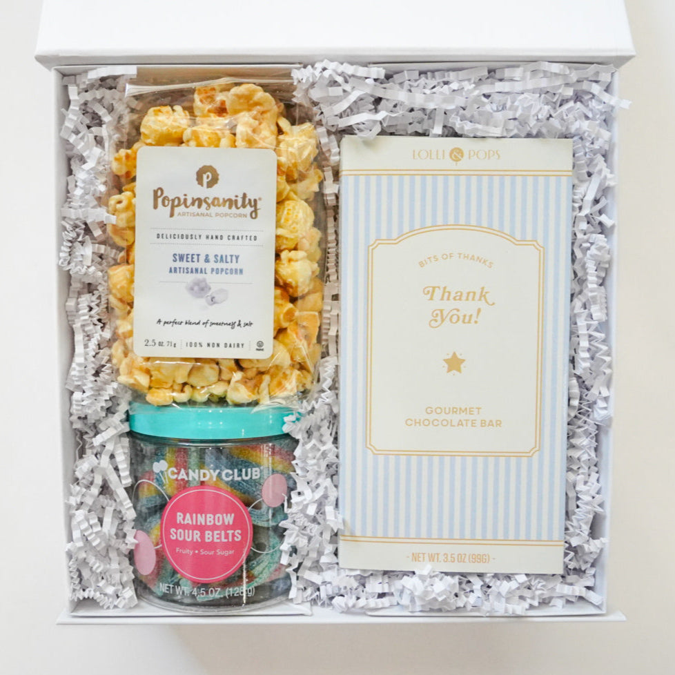Digital photograph of a white gourmet snack gift box featuring golden-brown Popinsanity popcorn, colorful Candy Club rainbow sour belts, and a blue-striped Lolli & Pops gourmet chocolate bar nestled in white shredded paper filler