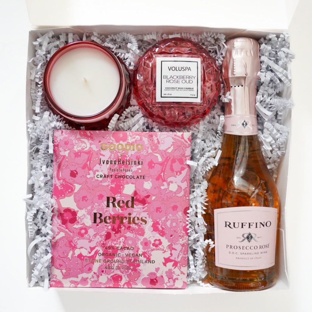 Photograph of a luxury gift box containing a bottle of Ruffino Prosecco Rose, a textured ruby Voluspa Blackberry Rose Oud candle, and a Goodio Ivana Helsinki craft chocolate bar with pink floral patterns nestled in white shredded paper