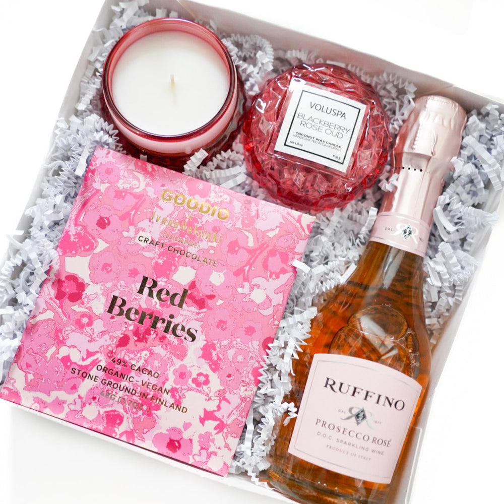 Digital photograph of a curated luxury gift box featuring a bottle of Ruffino Prosecco Rosé, a Voluspa Blackberry Rose Oud candle in a textured red glass jar, and a bar of Goodio Red Berries craft chocolate nestled in white crinkle paper filling