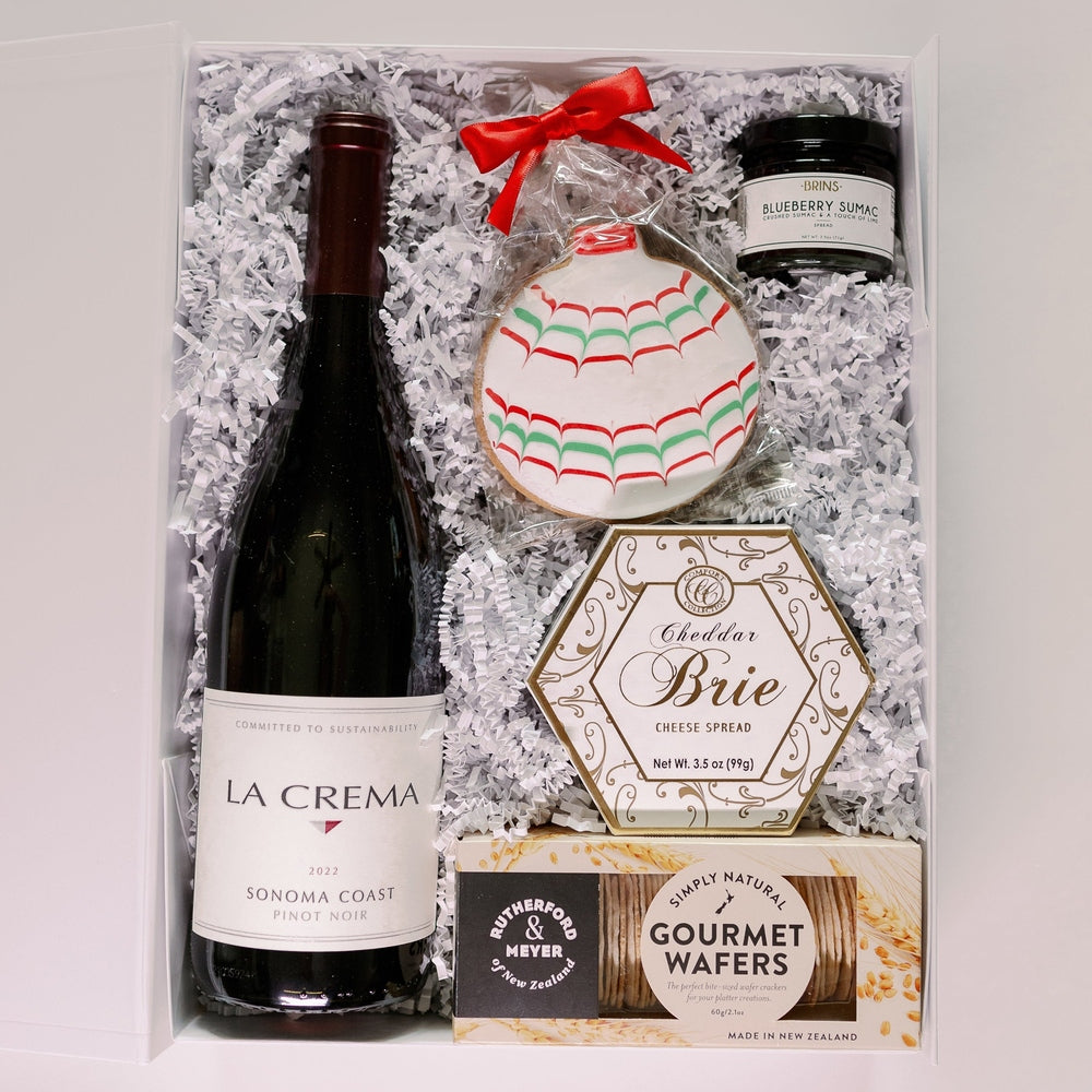 Photograph of a gourmet holiday gift box featuring a bottle of La Crema Sonoma Coast Pinot Noir, a festive ornament-shaped sugar cookie with a bright red bow, and artisan snacks like blueberry sumac spread nestled in white crinkle paper