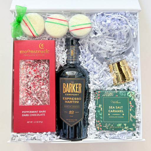 A curated gift box set containing a bottle of Barker Espresso Martini, a red box of Mannsbruch peppermint bark, a green box of sea salt caramel, three macarons, and a gold-wrapped item, arranged in white crinkle paper inside a white box.