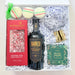 A curated gift box set containing a bottle of Barker Espresso Martini, a red box of Mannsbruch peppermint bark, a green box of sea salt caramel, three macarons, and a gold-wrapped item, arranged in white crinkle paper inside a white box.