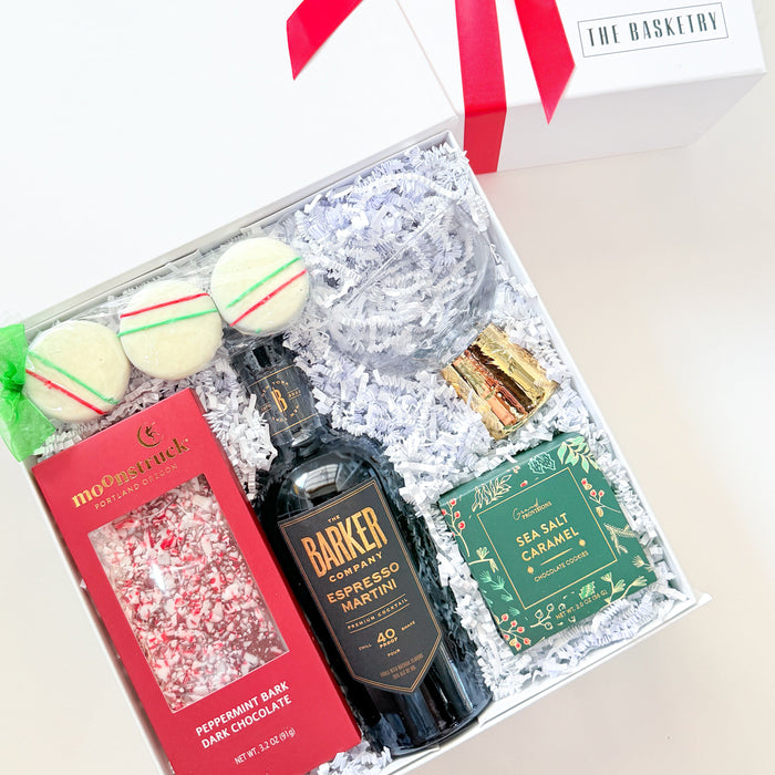 An open white gift box tied with a red ribbon, filled with assorted gift items on white crinkle paper. Visible contents include a dark bottle labeled Barkey's, a red box, a green package, macarons, and a clear glass tumbler.
