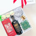 An open white gift box tied with a red ribbon, filled with assorted gift items on white crinkle paper. Visible contents include a dark bottle labeled Barkey's, a red box, a green package, macarons, and a clear glass tumbler.