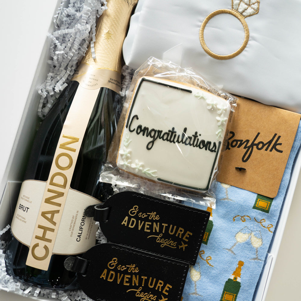Photograph of a wedding gift box featuring a Chandon California Brut bottle, a sugar cookie with black Congratulations script, and black leather luggage tags with gold foil adventure lettering next to a gold embroidered diamond ring and blue patterned socks